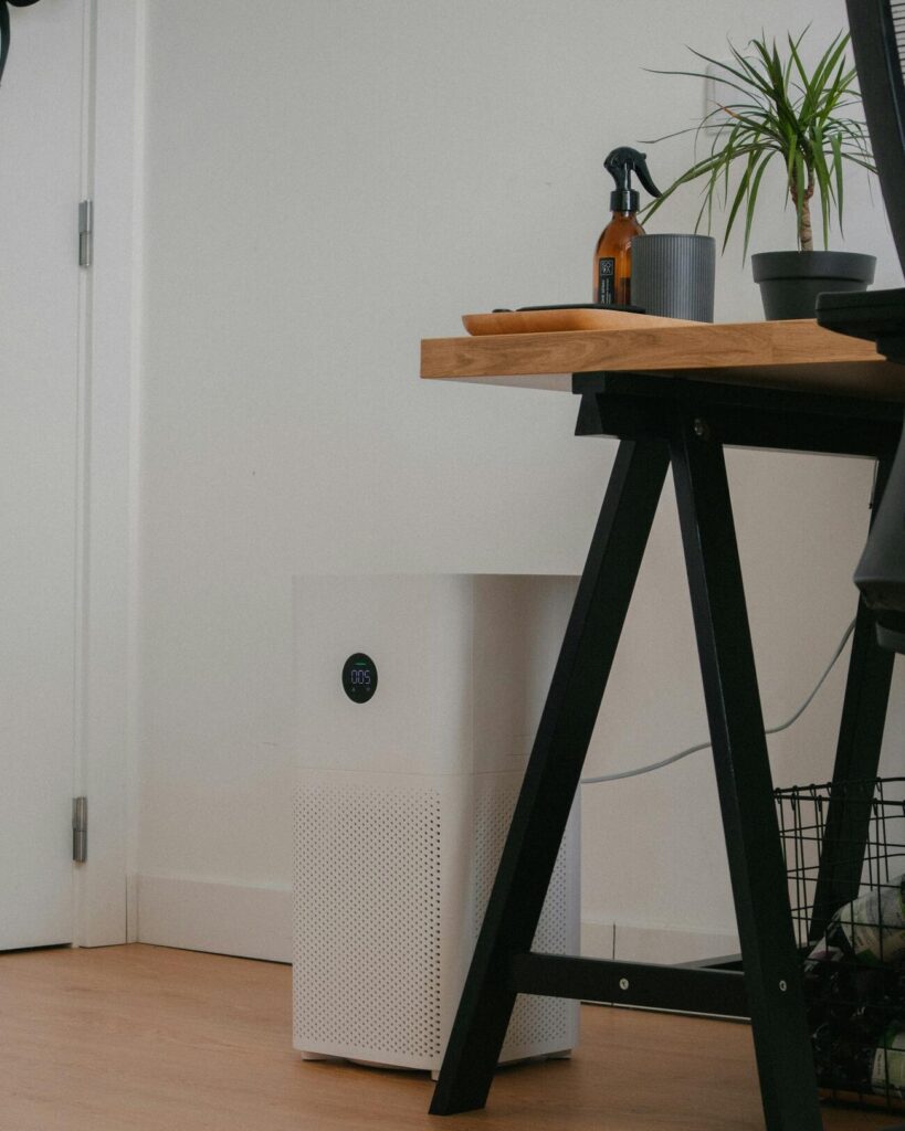 White air purifier on a wooden floor beneath a desk with potted plants and a spray bottle
