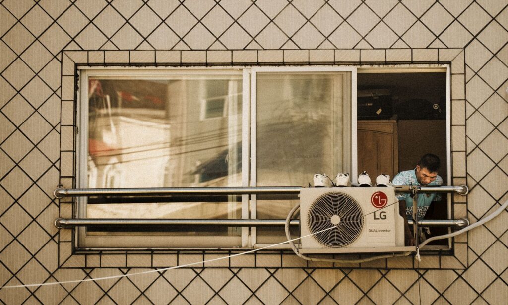 Man leaning out of a window beside an outdoor air conditioning unit mounted on a tiled building